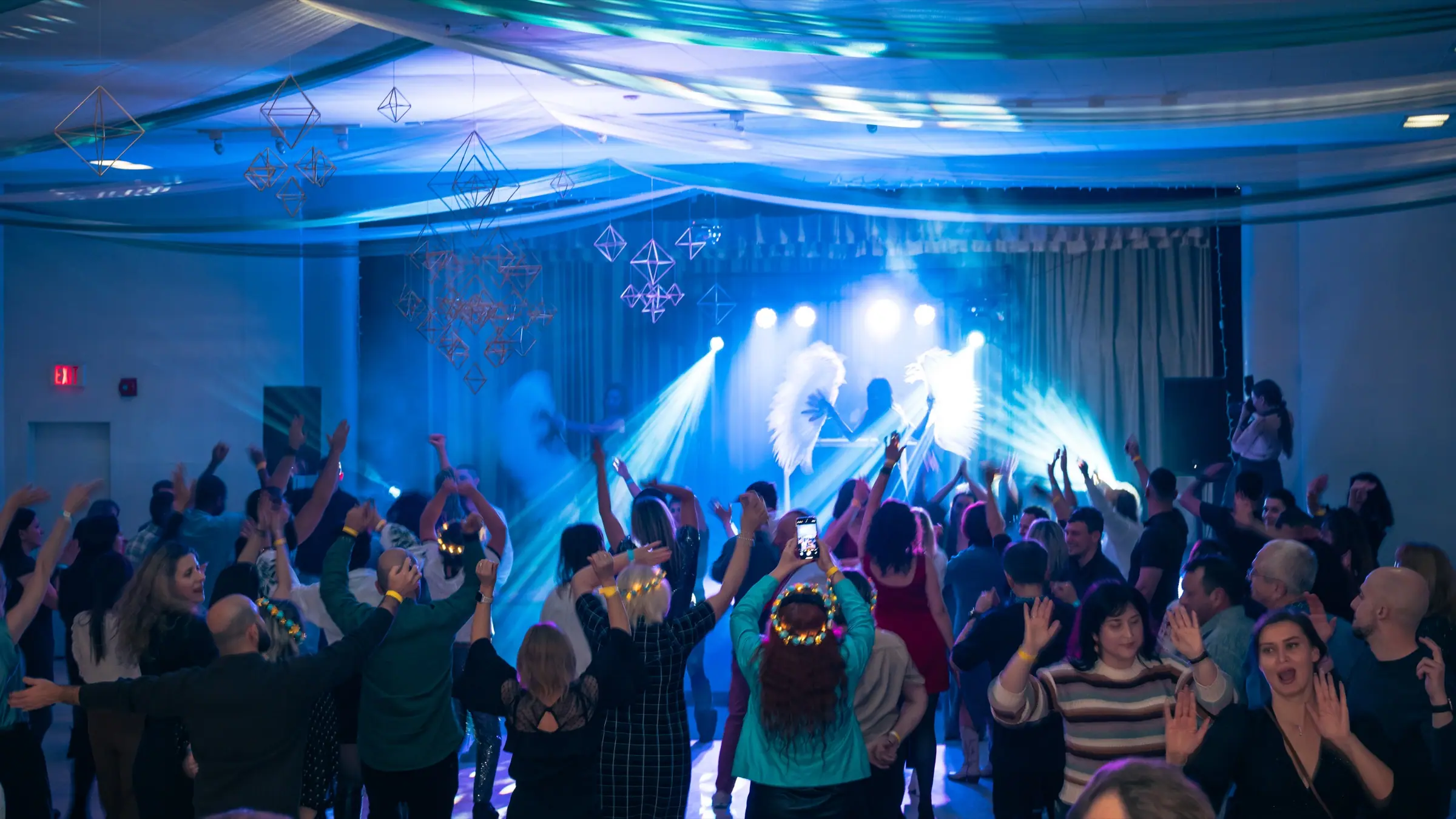 A full dance floor at Andriyivski Vechornytsi in Ottawa: guests with raised hands under blue stage light, straw ornaments hanging from the ceiling.